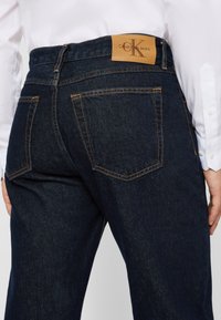 Tumedat sinised denim püksid kontrastsete oranžide õmblustega ja Calvin Kleini nahkplekk tagumisel taljal, millel on kaks tagumist taskut.