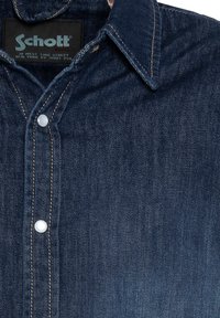 Camicia in denim blu scuro con colletto a punta, chiusura con bottoni bianchi e cuciture gialle a contrasto sul risvolto. Etichetta Schott visibile.