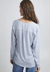 Blusa a maniche lunghe blu chiaro con vestibilità rilassata, scollatura rotonda e texture liscia. Il retro presenta un dettaglio con cucitura orizzontale.