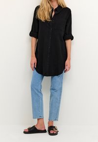 Chemise noire boutonnée avec manches retroussées, une poche ; associée à un jean bleu clair fuselé et des sandales noires avec une boucle dorée.