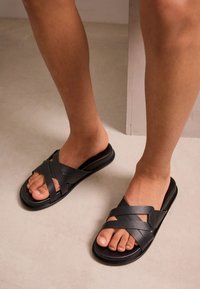 Schwarze Pantoletten mit überkreuzten Riemen, gefertigt aus glattem Material. Flaches Profil und offenes Design, das die Zehen betont und für Atmungsaktivität sorgt.