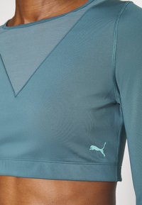 Teal sports crop top with a textured panel and a triangular design at the neckline. Features a small light blue Puma logo on the lower left.