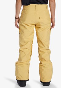 Geel sneeuwbroek met een hoge taille, cargozakken en een relaxte pasvorm. Voorzien van ritsen bij de enkels en een gladde, waterdichte textuur.