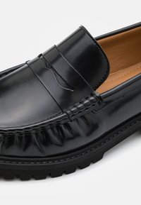 Mocassin en cuir noir avec une finition polie, présentant un design cousu, des détails texturés sur les côtés et une semelle en caoutchouc durable.