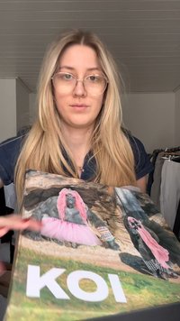 Joven con gafas sosteniendo una caja que muestra gorilas vestidos con tutús rosas y accesorios, con la palabra "KOI" en ella.