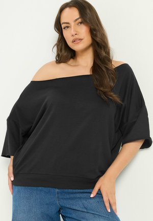 Kvinde med langt brunt hår iført en løs sort off-shoulder top og blå jeans, stående mod en ensfarvet lys væg.