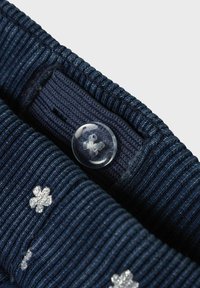 Cuello de tela ribeteada en color marino con un botón transparente que presenta un diseño floral. Bordado en plata de una flor visible en el cuello.