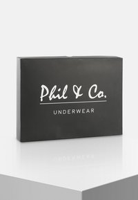 Zwarte rechthoekige doos met het label "Phil & Co. ONDERGOED" in witte schrijfletters, met een gladde matte textuur. Geplaatst op een reflecterend oppervlak.
