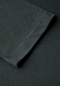 Tessuto di cotone verde scuro con una texture liscia; presenta un bordo del polsino cucito e una leggera costina, che indicano un design aderente.