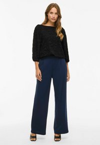 Blusa nera a maniche lunghe con texture abbinata a pantaloni larghi blu navy. L'outfit è completato da sandali neri con tacco.