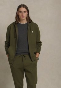 Σκούρο πράσινο φούτερ με λογότυπο, συνδυασμένο με γκρι φούτερ και ταιριαστά παντελόνια φόρμας. Το σύνολο έχει άνετη εφαρμογή και τσέπες.