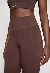 Mujer vistiendo leggings de cintura alta marrones y un sujetador deportivo a juego con costuras visibles y el logo de Salomon en el sujetador.