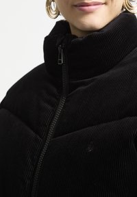 Musta corduroy-takki, jossa on korkea kaulus ja vetoketjusulku. Takissa on paksu tekstuuri ja hienovarainen logo rinnassa.