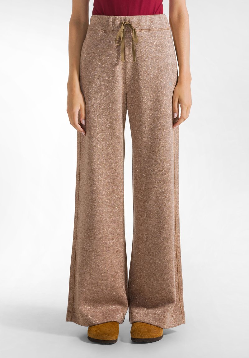 Bruine, wijde broek met een gestructureerde, glinsterende afwerking. Voorzien van een elastische tailleband en verstelbaar trekkoord. Gecombineerd met gele schoenen.