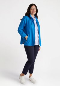 Niebieska wodoodporna kurtka z zamkiem błyskawicznym z przodu, kieszeniami po bokach i regulowanym kapturem, w połączeniu z granatowymi spodniami i białymi trampkami.