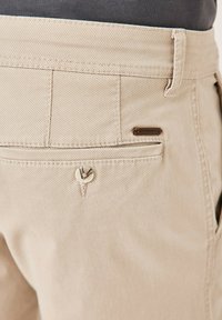 Beige Hosen aus strukturiertem Stoff, mit einer Gesäßtasche, die mit einem Knopf gesichert ist, und einem kleinen braunen Etikett.