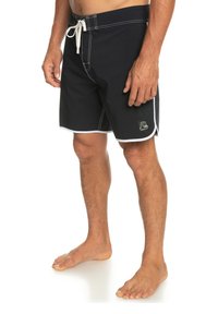 Man wearing black swim trunks with white trim and drawstring, standing barefoot against a white background.