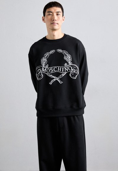 Svart sweatshirt med långa ärmar, som har en vit grafisk design av lagerblad och "MOSCHINO"-logotypen. Kombineras med svarta byxor.