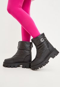 Botine negre cu un upper texturat și pufos și un toc gros, cu striații. Prezintă un fermoar pe lateral și un logo circular pe partea exterioară a tălpii.