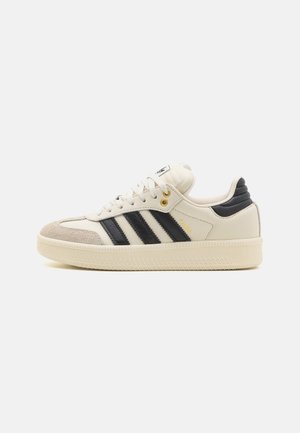 Kremowo-czarne tenisówki Adidas Samba z zamszowym noskiem, czarnymi paskami, złotymi oczkami i teksturowaną gumową podeszwą na białym tle.