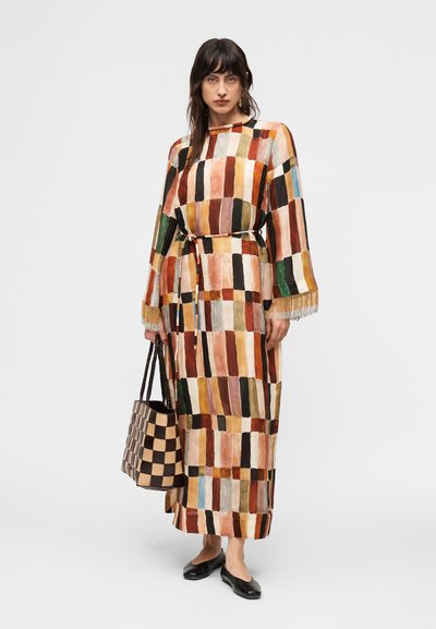 Γυναίκα που φοράει πολύχρωμο μακρύ καρό φόρεμα με μανίκια με φράντζες, κρατώντας μια μεγάλη καφέ και μπεζ καρό τσάντα, με μαύρα επίπεδα παπούτσια.