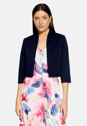 Femme portant un blazer court bleu marine sur une robe portefeuille fleurie rose, bleue et blanche à manches trois-quarts.