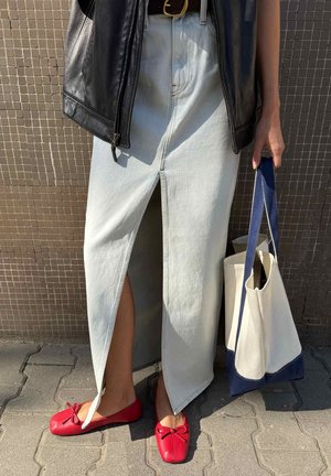 Isik, kes kannab heledeniimseid seelikut eesmise lõhikuga, musta nahkjopet, punaseid kingi, hoides valget ja sinist käekotti plaatidel kõnniteel.