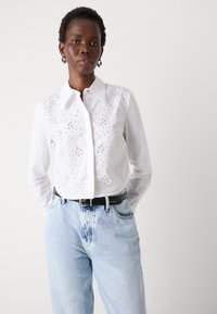 Camicia bianca con bottoni, caratterizzata da un frontale texturizzato con borchie metalliche, abbinata a jeans di denim blu chiaro e a una cintura nera.
