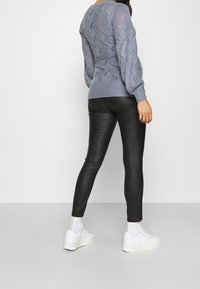 Mujer vestida con un suéter de punto lavanda con cinturón, pantalones de cuero negros, calcetines blancos y zapatillas blancas de suela gruesa, de pie contra un fondo blanco.