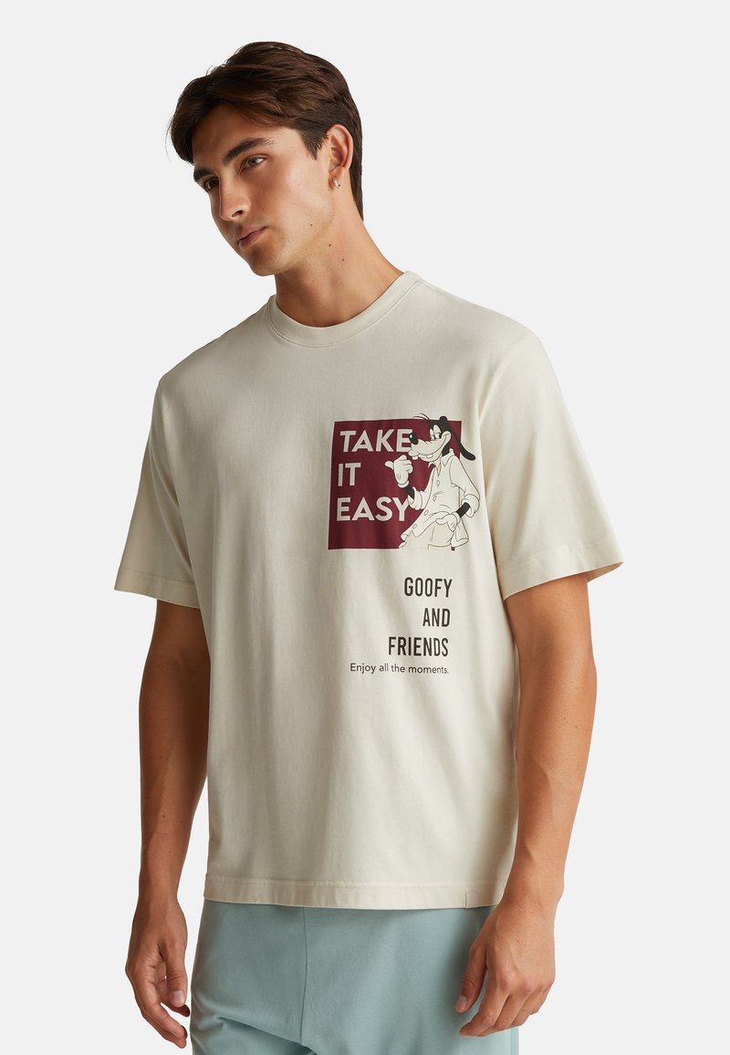 Mladý muž má na sobě krémové tričko s grafikou Goofy a textem "Ber to s klidem, Goofy a přátelé, Užij si všechny chvíle."