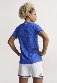 Modrá sportovní dres s texturovaným vzorem listů, zlatými akcenty a bílými šortkami. Má kulatý výstřih a krátké rukávy.