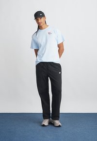 Gai zils Nike T-krekls ar logo, melnas brīvā stila bikses un pelēki sporta apavi; modelis ar melnu beisbola cepuri ar logo.