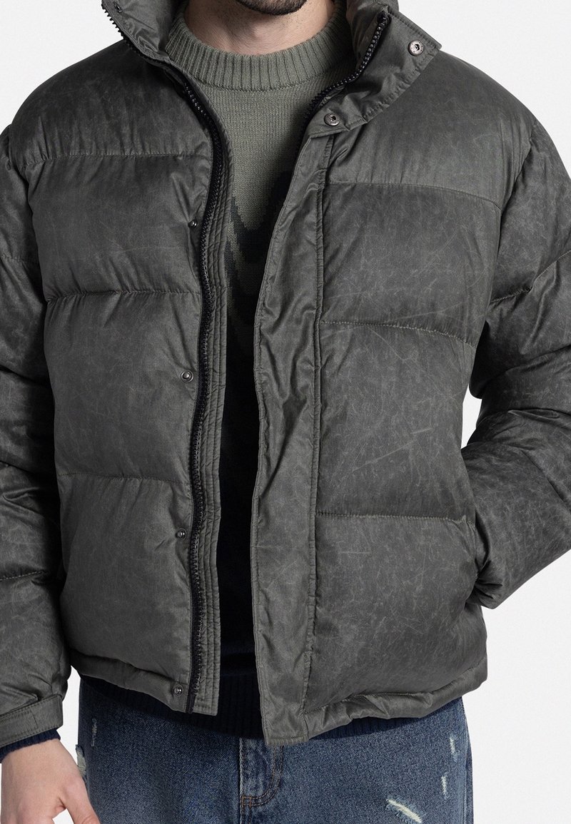 Uomo che indossa una giacca imbottita grigio scuro sopra un maglione di cotone verde oliva e jeans blu strappati, con le mani nelle tasche della giacca.