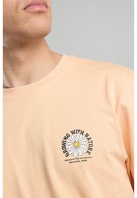 Light peach cotton t-shirt featuring a round graphic print with a daisy and text "GROWING WITH NATURE" centered on the chest.