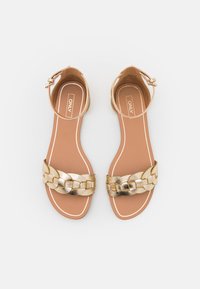 Gold strappy sandals with a braided metallic strap, a flat sole, and a buckled ankle strap. Smooth texture with a brown footbed and white trim.