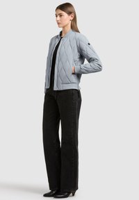 Femme portant une veste bomber matelassée bleu clair, un haut noir, un jean noir à jambes larges et des bottes à talons noires, debout avec une main dans la poche.