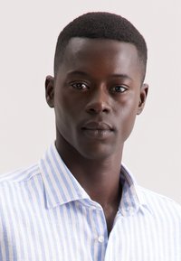 Camicia a righe blu e bianche con bottoni, colletto corto, tessuto leggero, bottoni in vista e vestibilità rilassata. Primo piano di una persona che la indossa.