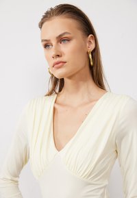 Light yellow dress with long sleeves, V-neck, and gathered detailing at the bust. Model wears large gold hoop earrings. Smooth texture.