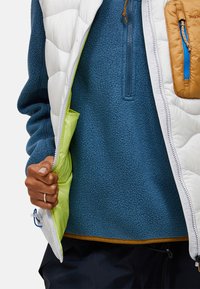 Vit pufferväst med en ljusgrön foder över en blå fleece-tröja, med en brun ficka med dragkedja i blått.