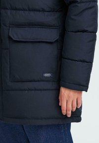 En manns hånd ved siden av en stor svart polstret lomme med klaff, liten etikett og synlige blå denimbukser.