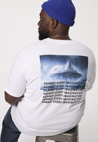 Hvid bomulds t-shirt med et grafisk motiv af et futuristisk landskab på ryggen, med gentaget tekst "IMOD HVER FANTASI" ovenfor.