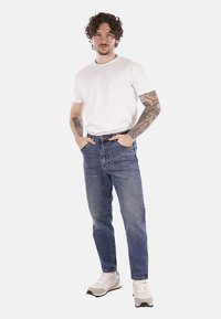 T-shirt bianca in cotone, jeans in denim blu e sneakers beige. Il modello sta in piedi con le mani nelle tasche, mostrando il taglio casual e i dettagli di design.
