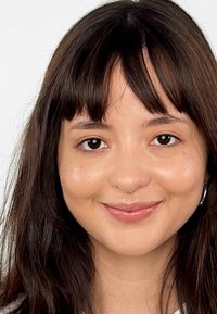 Cheveux châtain foncé de longueur moyenne, teint de peau doux et maquillage naturel. Les traits remarquables incluent un léger sourire et des boucles d'oreilles créoles.