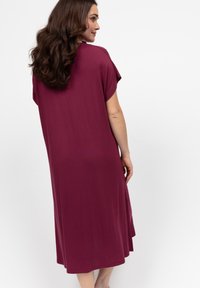 Kobieta z długimi falującymi włosami, ubrana w luźną, krótką sukienkę midi w kolorze burgundowym, stojąca lekko zwrócona bokiem na białym tle.