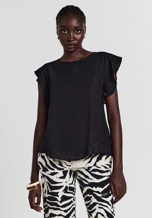 Donna con capelli intrecciati che indossa un top smanicato nero con maniche a volant e pantaloni a stampa zebra, accessoriata con bracciali sul polso sinistro.