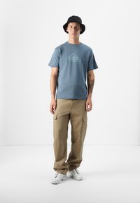 En ung mann står i et minimalistisk studio, iført en blå t-skjorte, beige cargo-bukser og svart bøttehatt, med et fokusert uttrykk.