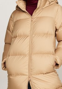 Beige gewatteerde jas met capuchon, voorzien van een ritssluiting, geïsoleerd ontwerp en een doorgestikt patroon. Gelaagd buitenmateriaal en zijzakken.