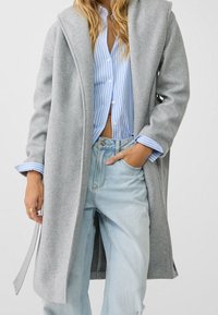 Mujer llevando una camisa de botones a rayas azul claro, unos jeans azul claro y un abrigo largo gris abierto, con una mano en el bolsillo.