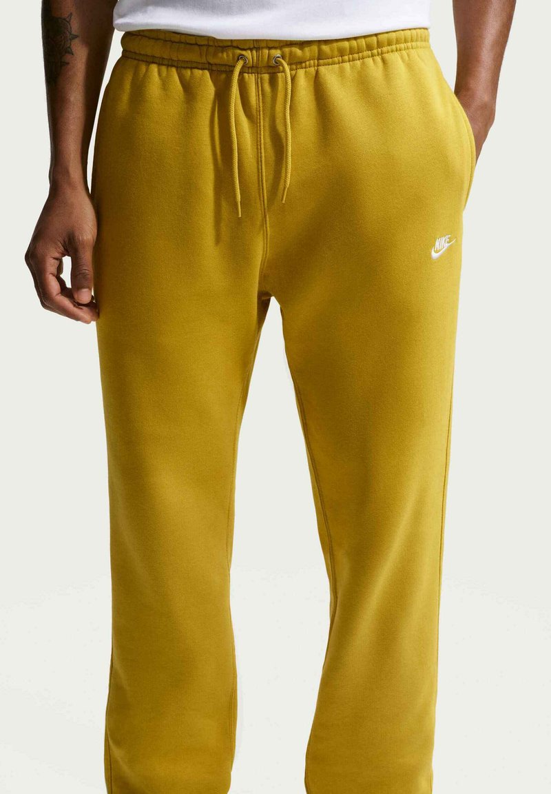 Homme portant un pantalon de survêtement Nike jaune moutarde avec taille à cordon et logo Nike blanc sur la cuisse droite, mains dans les poches et le long du corps.