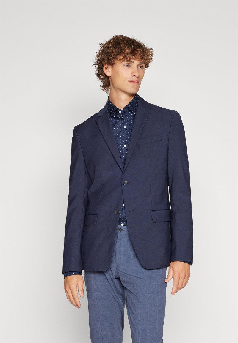 Navy blazer with notch lapels, two front pockets, and single button closure, paired with a dark polka dot shirt and light blue checked pants.